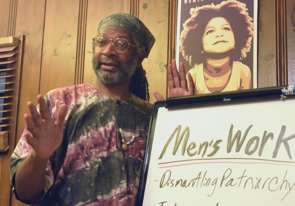 Promotional Portrait of Facilitator Ukumbwa Sauti, M.Ed. at the Democracy Center, Cambridge, MA USAmerica, gesturing, next to a white board that says, "Men's Work, Dismantling Patriarchy"