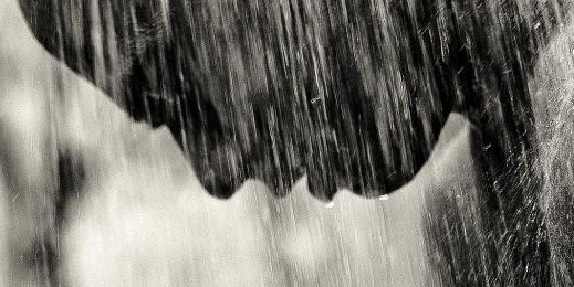 portrait from side of man hanging his head as water pours over him as if in heavy rainshower, close-up in black and white