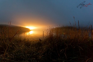 Low key, wide angle shot of water surrounded by marsh and hills at sunrise, a warm yellow sun just at the horizon