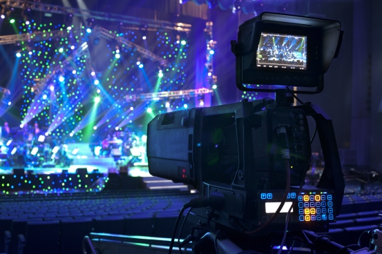 image of television camera in foreground with concert stage in background, coolly lit with numerous lighting units